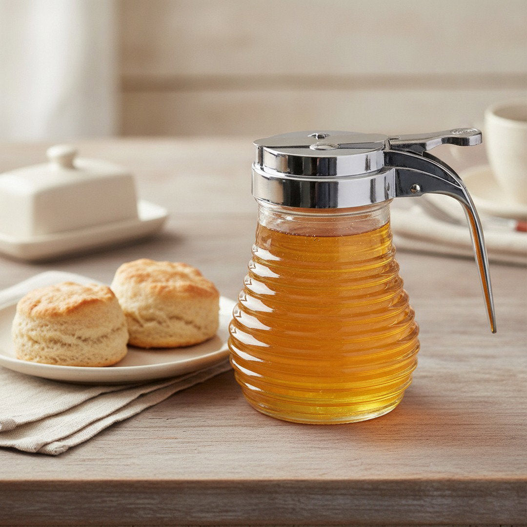 Beehive glass syrup dispenser filled with golden maple syrup beside warm biscuits on a softly lit breakfast table.