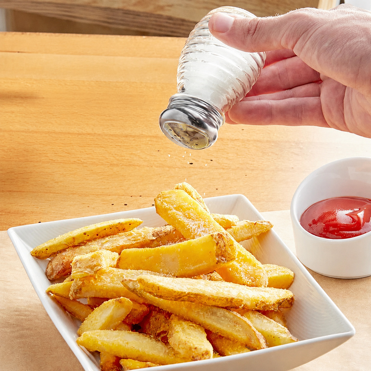 Hand using a diner-style beehive glass salt shaker to season golden French fries beside a cup of ketchup.