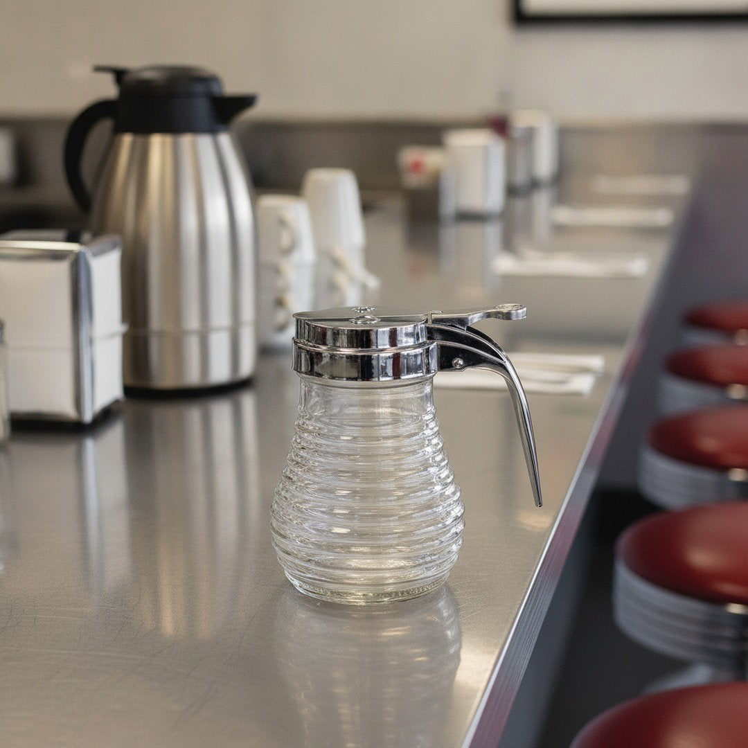 Empty ribbed glass beehive syrup dispenser with chrome pour top displayed on a stainless-steel diner counter.